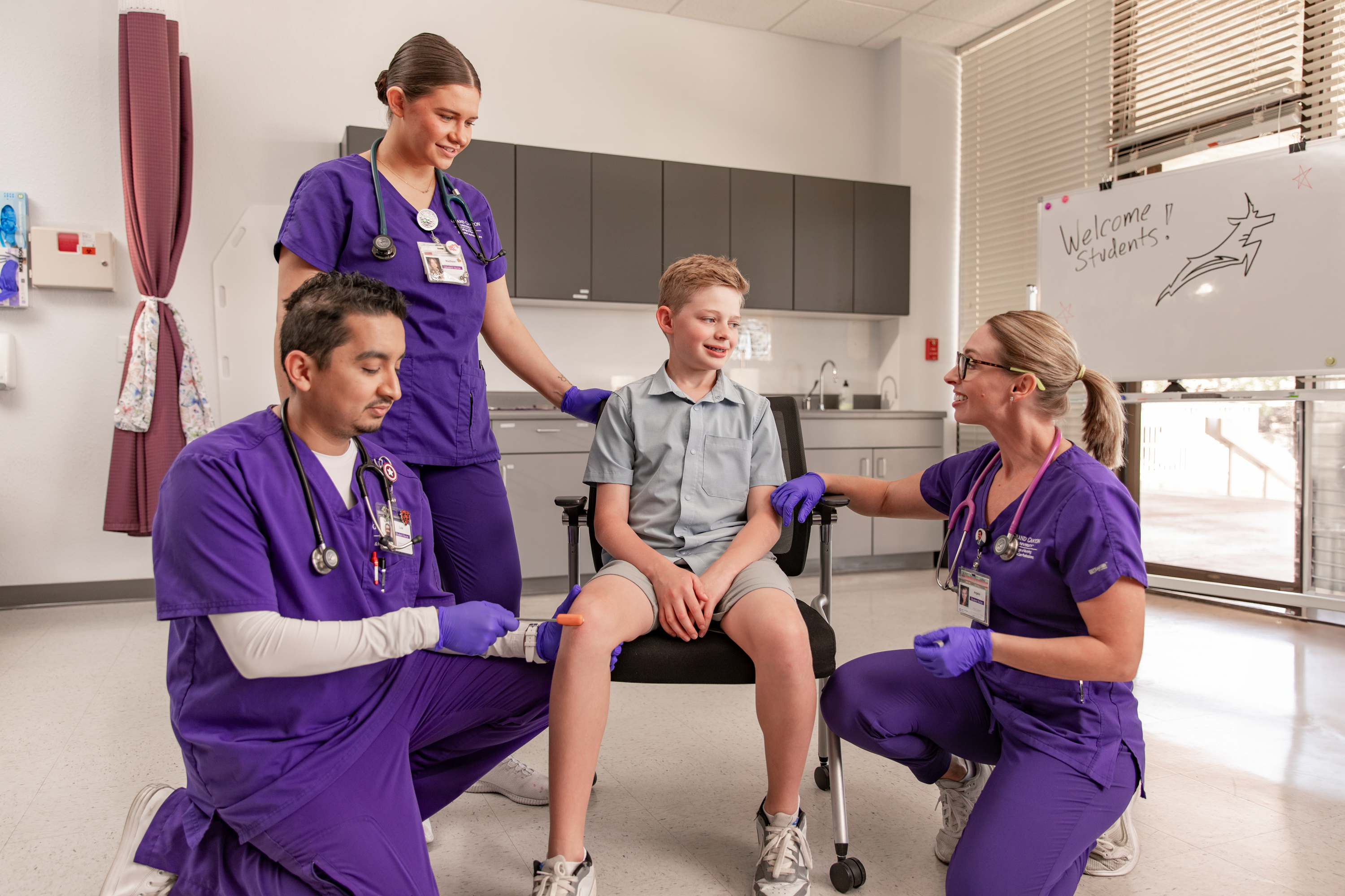 Three nurses in purple slacks, helping a young patient in a chair